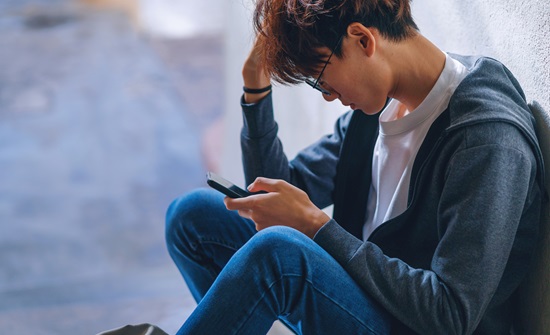 Teen boy sitting outside on the ground with his legs crossed and looking at his phone