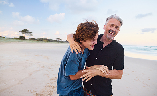 father and teen son walking on beach and hugging and laughing