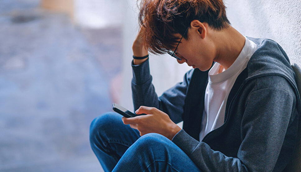 teen boy sitting on ground outside with legs crossed, looking at phone, deep in thought